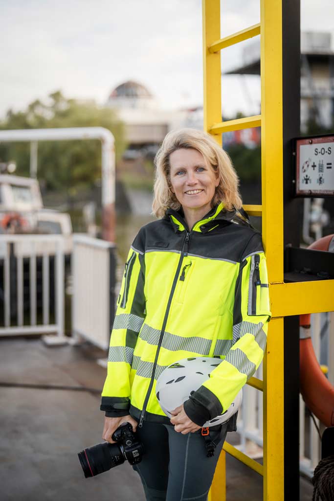 Britta Hilpert: Portraitbild mit Kamera, Warnjacke und Helm unter dem Arm